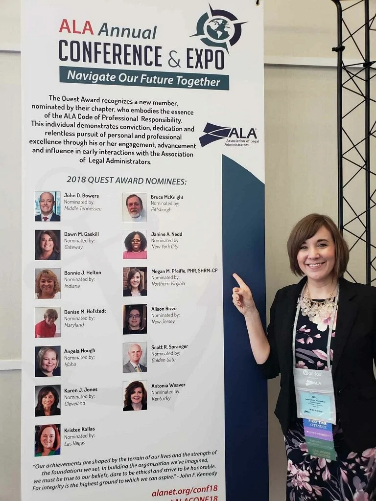 A woman standing next to a conference poster for the ALA Annual Conference & Expo. She is smiling and pointing at the poster, wearing a black blazer, floral dress, and conference badge. The poster lists 2018 Quest Award nominees, includes the ALA log