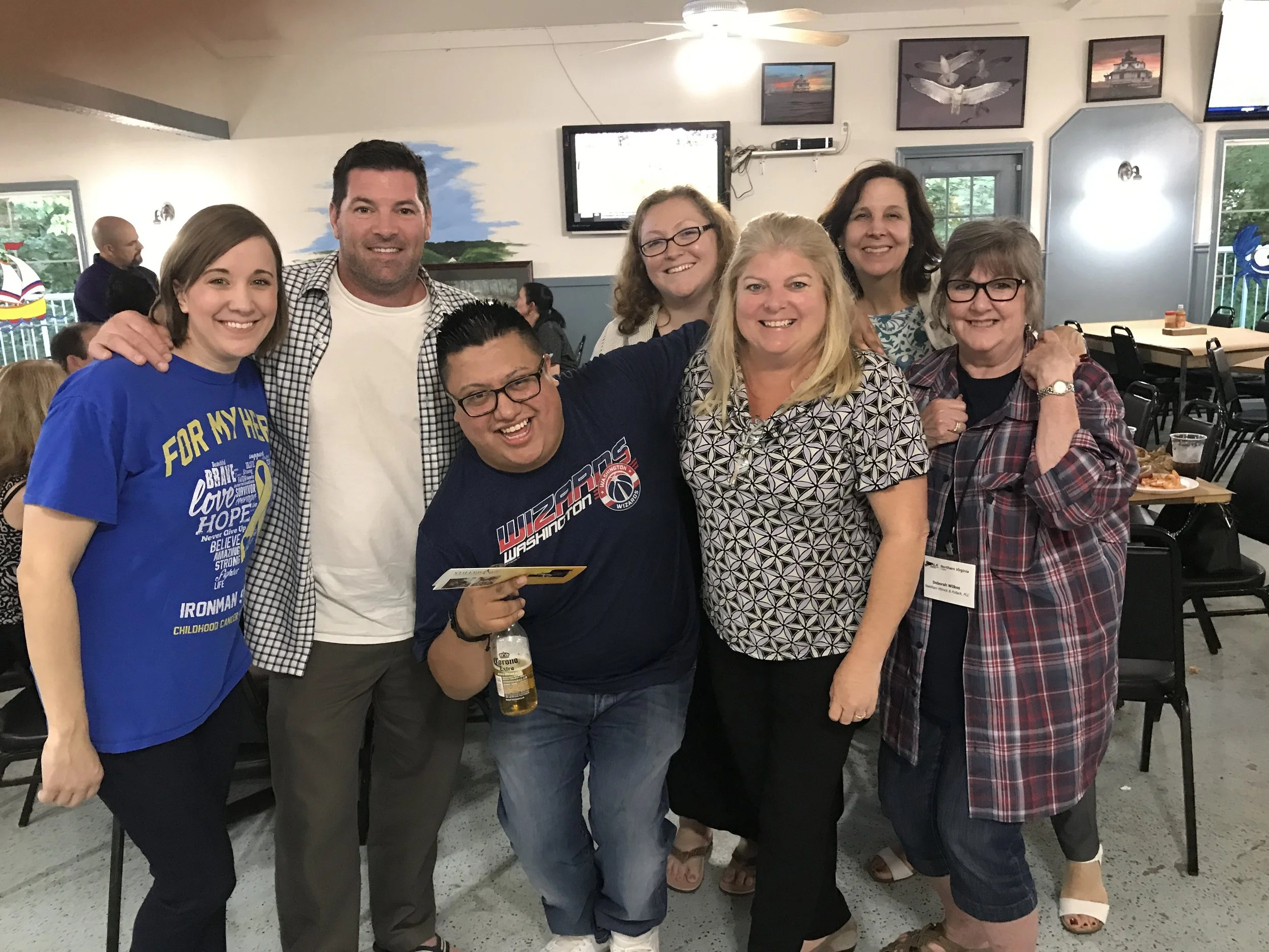 A group of seven people smiling and posing together indoors, with a woman in a blue T-shirt standing at the left and a woman in a plaid shirt at the right, all standing close with some holding drinks, in a room with wall art including paintings of sh