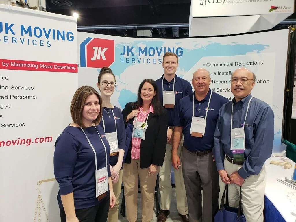 Six people standing in front of a JK Moving Services booth at an event or trade show, smiling at the camera.
