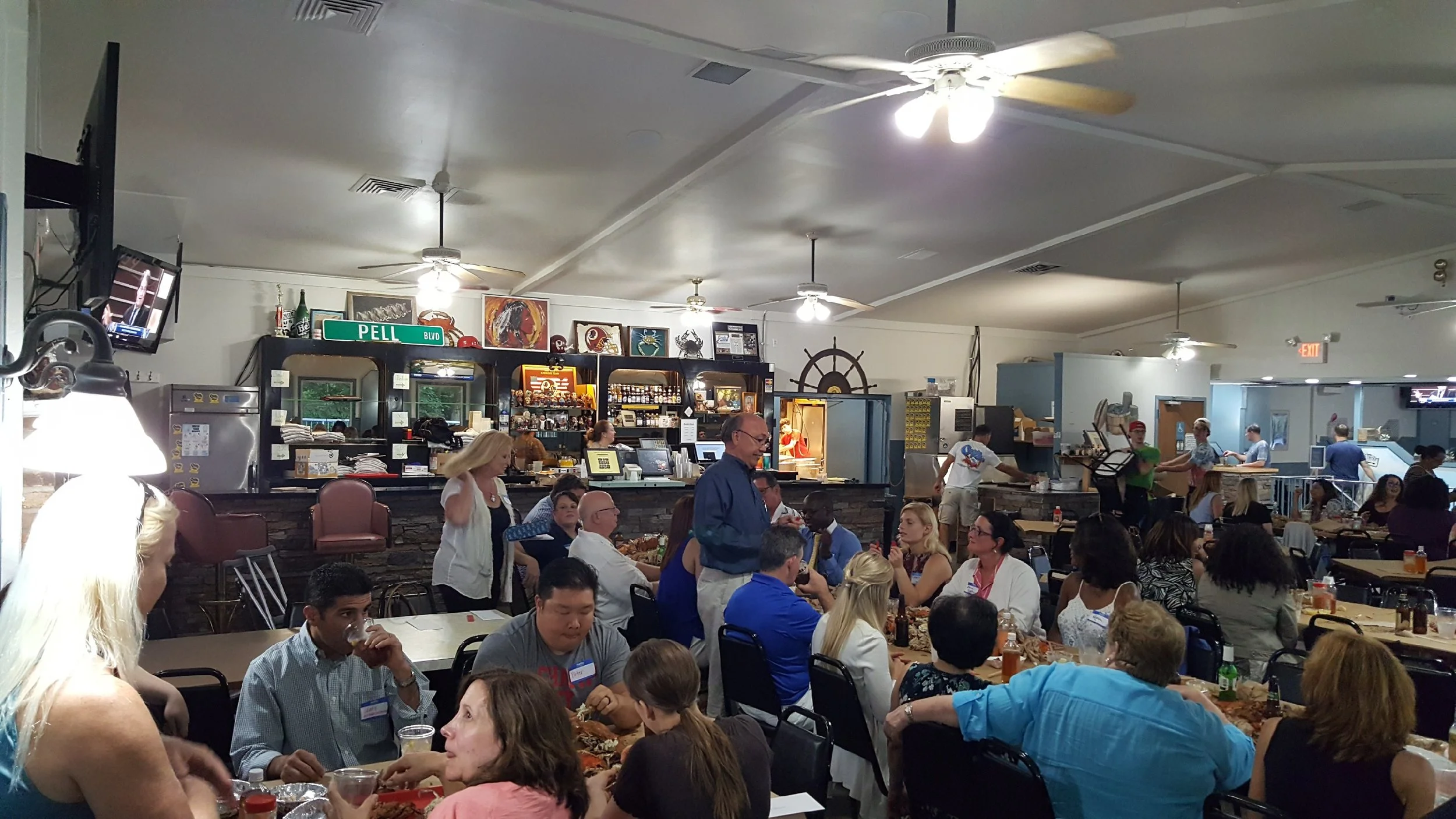 A crowded restaurant with people dining, servers serving food, a counter in the background, and a television on the wall.