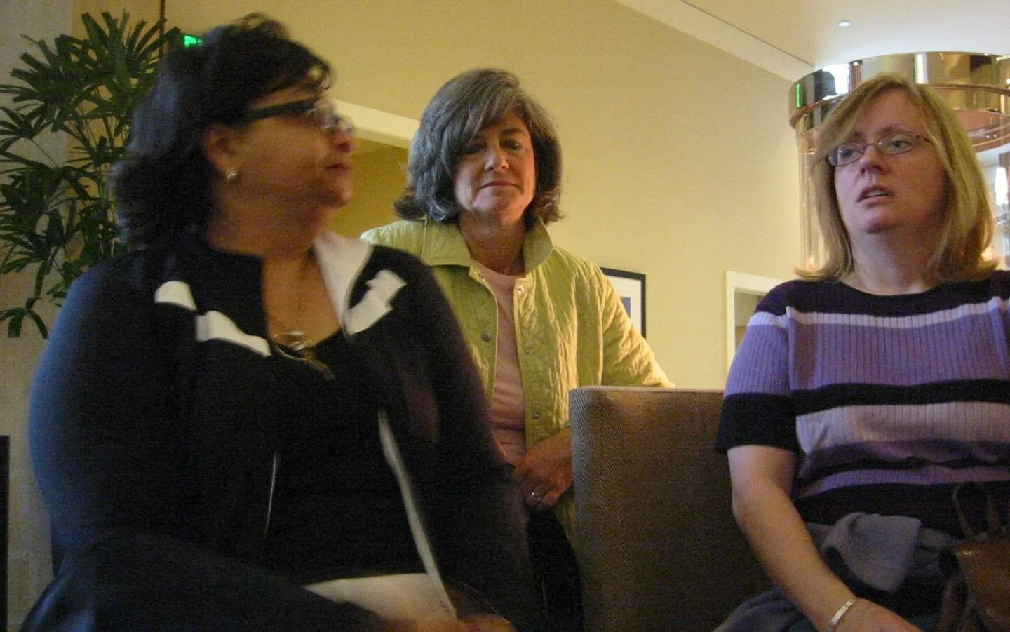 Three women sitting in a room, engaged in conversation, with a plant and framed pictures on the wall behind them.