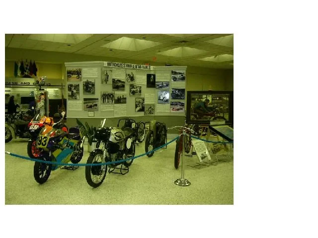 Display of vintage motorcycles and related memorabilia in a museum, with historical photographs on the wall.