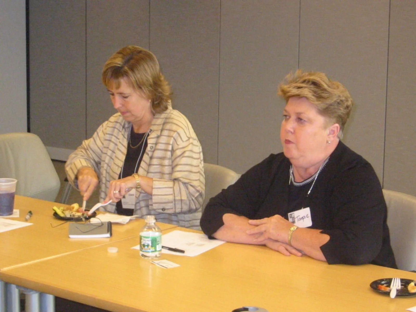 Two women sitting at a conference table, one slicing food on a plate, the other looking to her right, both with drink and papers on the table.