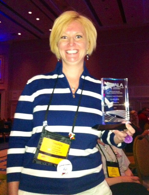 Woman with blonde hair smiling and holding a glass award at an indoor event conference, wearing a blue and white striped shirt and conference badge.