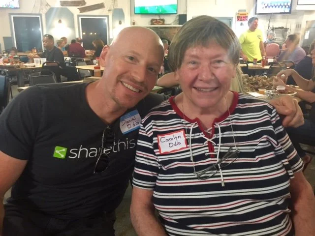 Two people smiling and sitting together at a restaurant or bar, with other patrons and television screens in the background. They are wearing name tags and casual clothing.