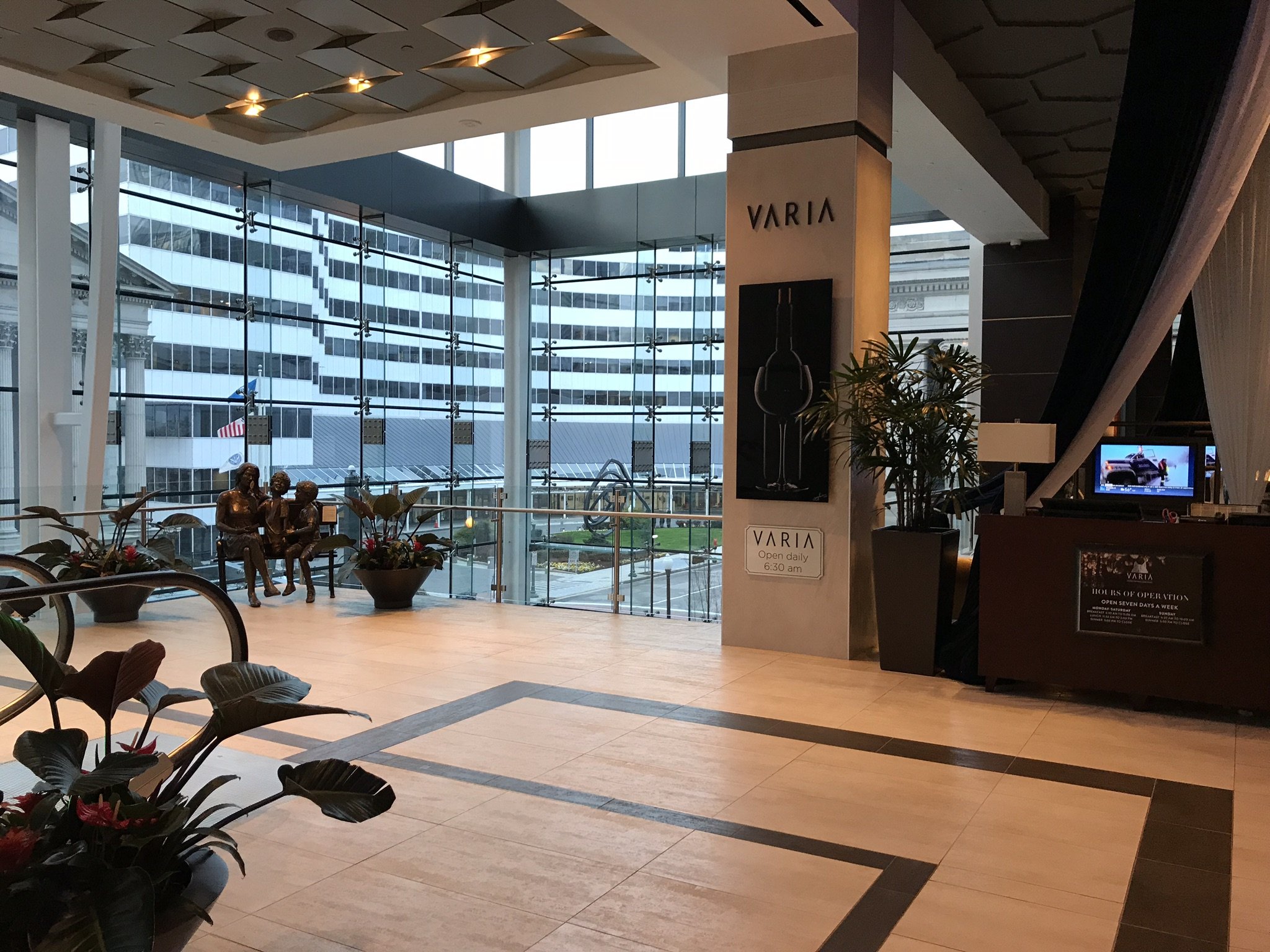 Interior of a modern shopping mall with a large glass window showing a city skyscraper outside. There are decorative plants and a bronze sculpture of children in the foreground. A sign reads 'VARIA,' indicating a store or brand, with operating hours 