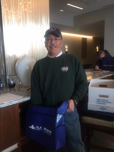A man wearing a green sweatshirt with a poker logo, baseball cap, and glasses standing indoors, holding a blue Virginia bag. There are decorative curtains, a large vase, and a box on a counter behind him.