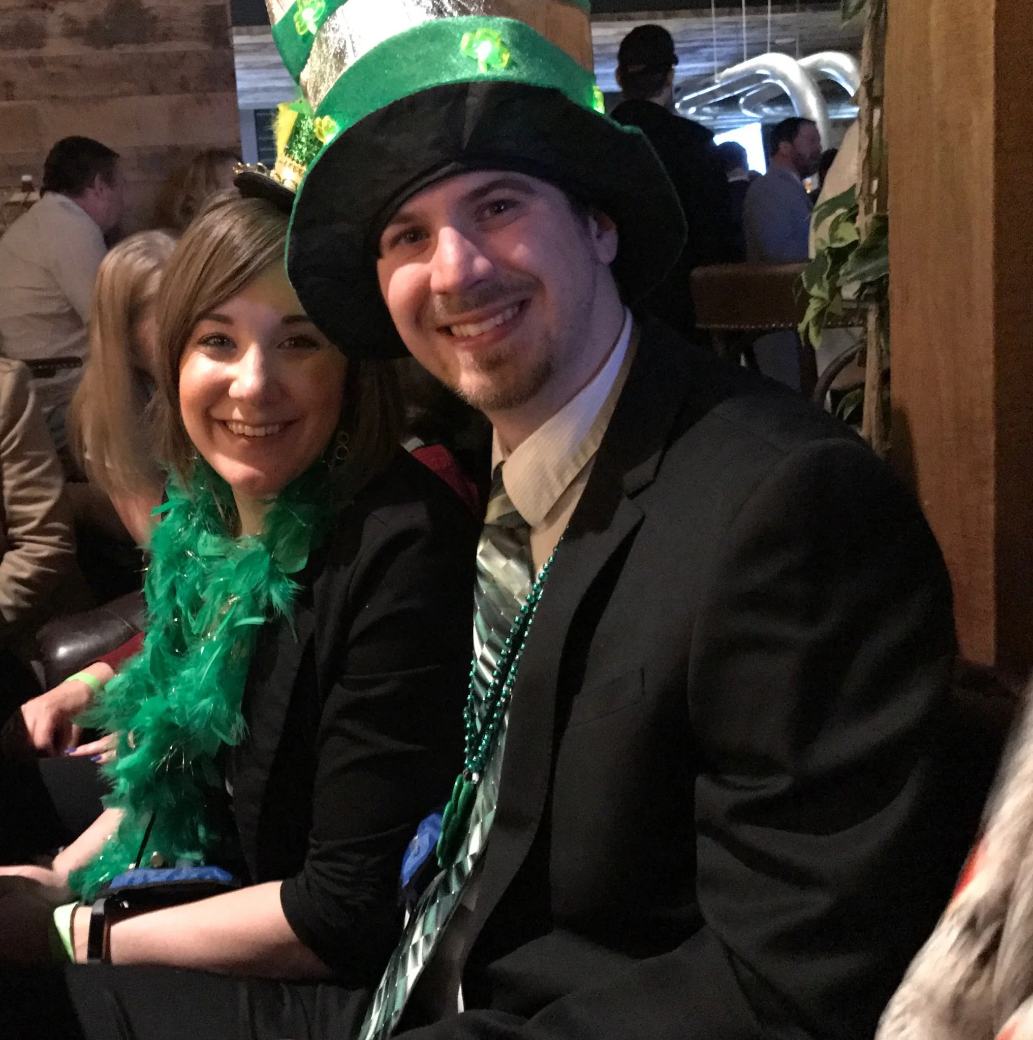 Young man wearing a large green St. Patrick's Day hat and green beads, smiling at a festive gathering.
