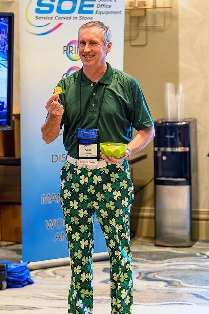 A man smiling and holding a yellow coin and a green bowl at a conference or event, standing in front of a booth with a sign.