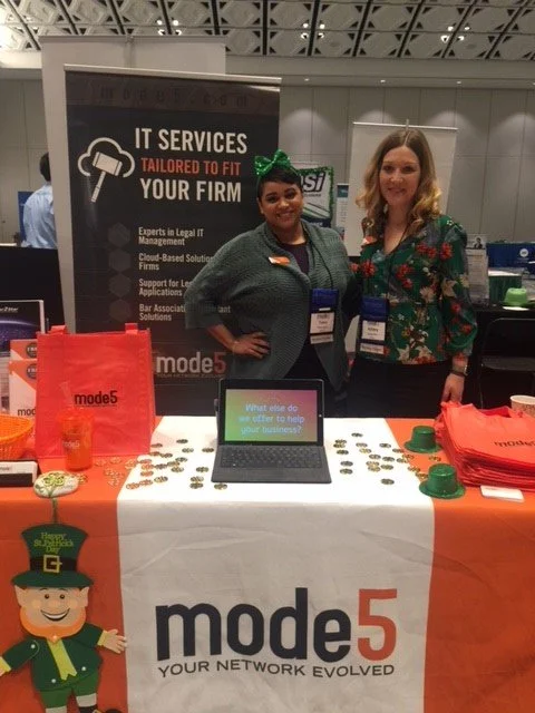 Two women at a technology booth, with a banner for IT services and a laptop displaying a question, surrounded by promotional materials and St. Patrick's Day decorations.