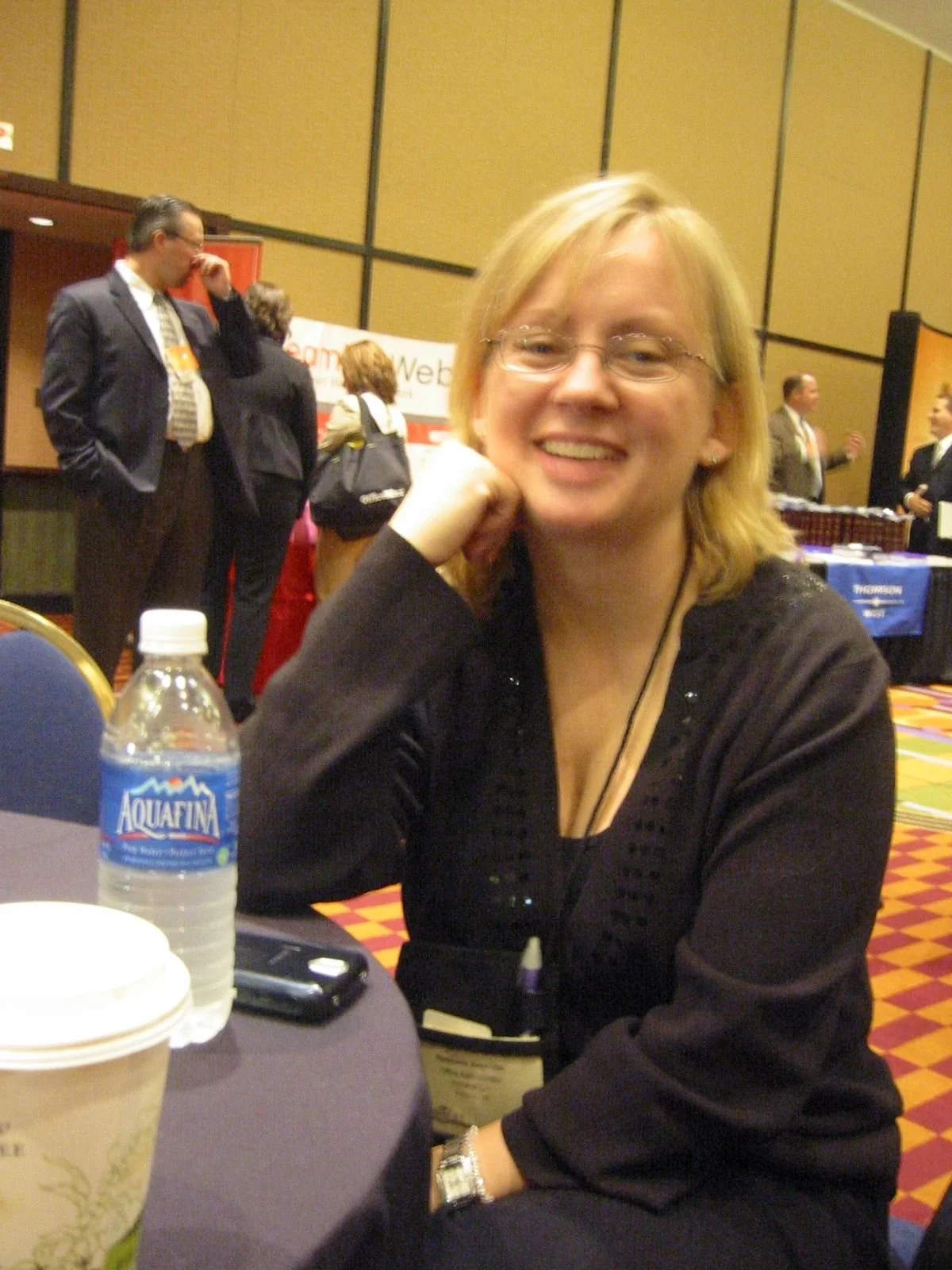 A smiling woman with blonde hair and glasses sitting at a table in a conference or event room. In the background, people are talking near a booth with a banner.