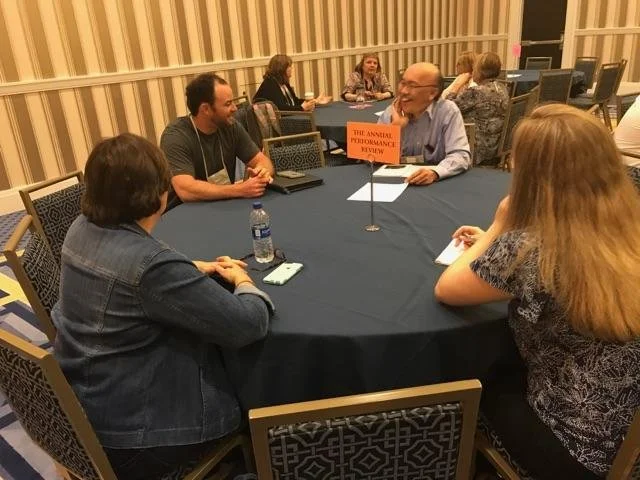 People sitting around a round table at a conference, engaged in conversation, with a sign on the table that reads 'The Annual Performance Review.'