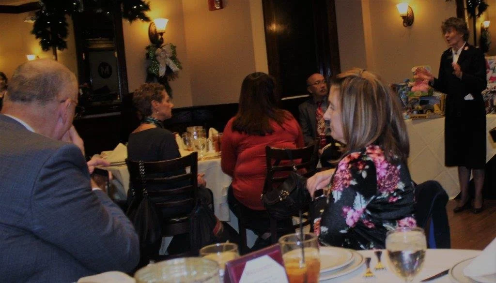 A woman dressed in a navy blue uniform speaking to a group of people at a dinner event in a restaurant decorated with holiday wreaths and lights.