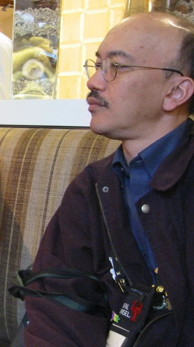 A man with glasses and a mustache sitting on a striped upholstered bench, wearing a dark jacket and blue shirt, with party accessories hanging from his jacket.