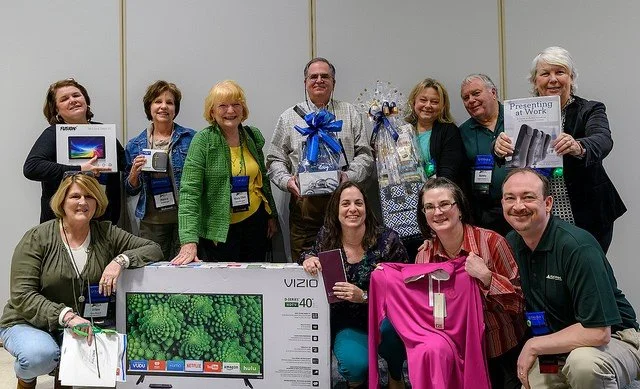 Group of ten people at an event, holding various products and gifts, standing behind a large TV box, with some giving thumbs up.