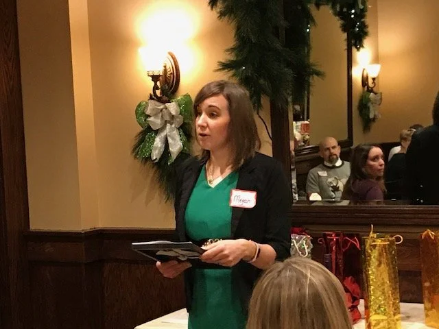 A woman with a name tag that says 'Megan' is standing and speaking to a group in a festive setting, decorated with a Christmas wreath and garland. There are gift bags on the table, and people seated in the background.