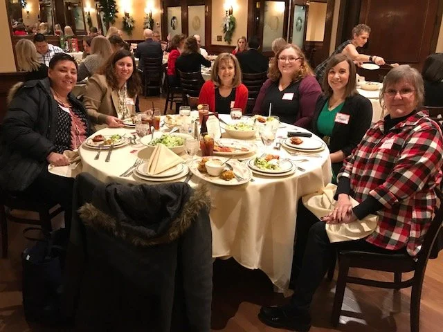 Group of six women seated around a round table in a restaurant, with plates and drinks, smiling at the camera.