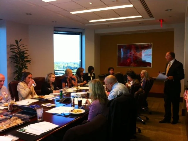 Business meeting in a conference room with adults sitting around a long table, a man standing with papers, and a window showing a city view.