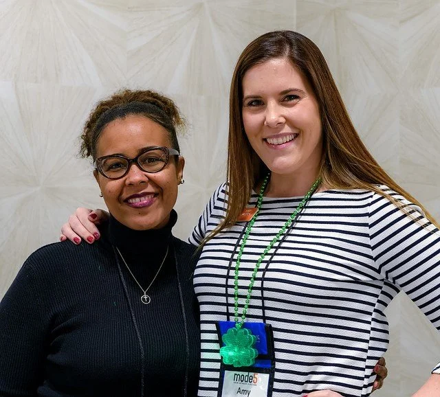 Two women smiling and posing together in front of a light-colored wall. One woman has short curly hair, glasses, and is wearing a black turtleneck. The other woman has long straight hair, is wearing a striped shirt, and has a green lanyard with a lar