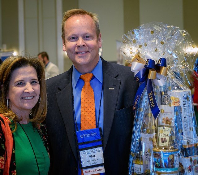 A man and woman smiling at an indoor event, with the man holding a large gift basket wrapped in clear plastic with blue, gold, and white ribbons.