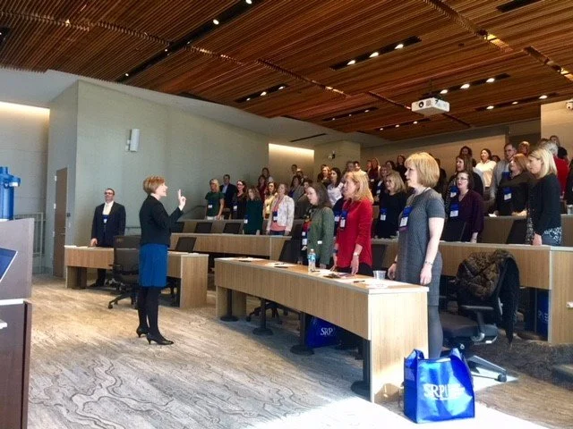 A woman is speaking or presenting to an audience that is standing and facing her in a conference or seminar room.