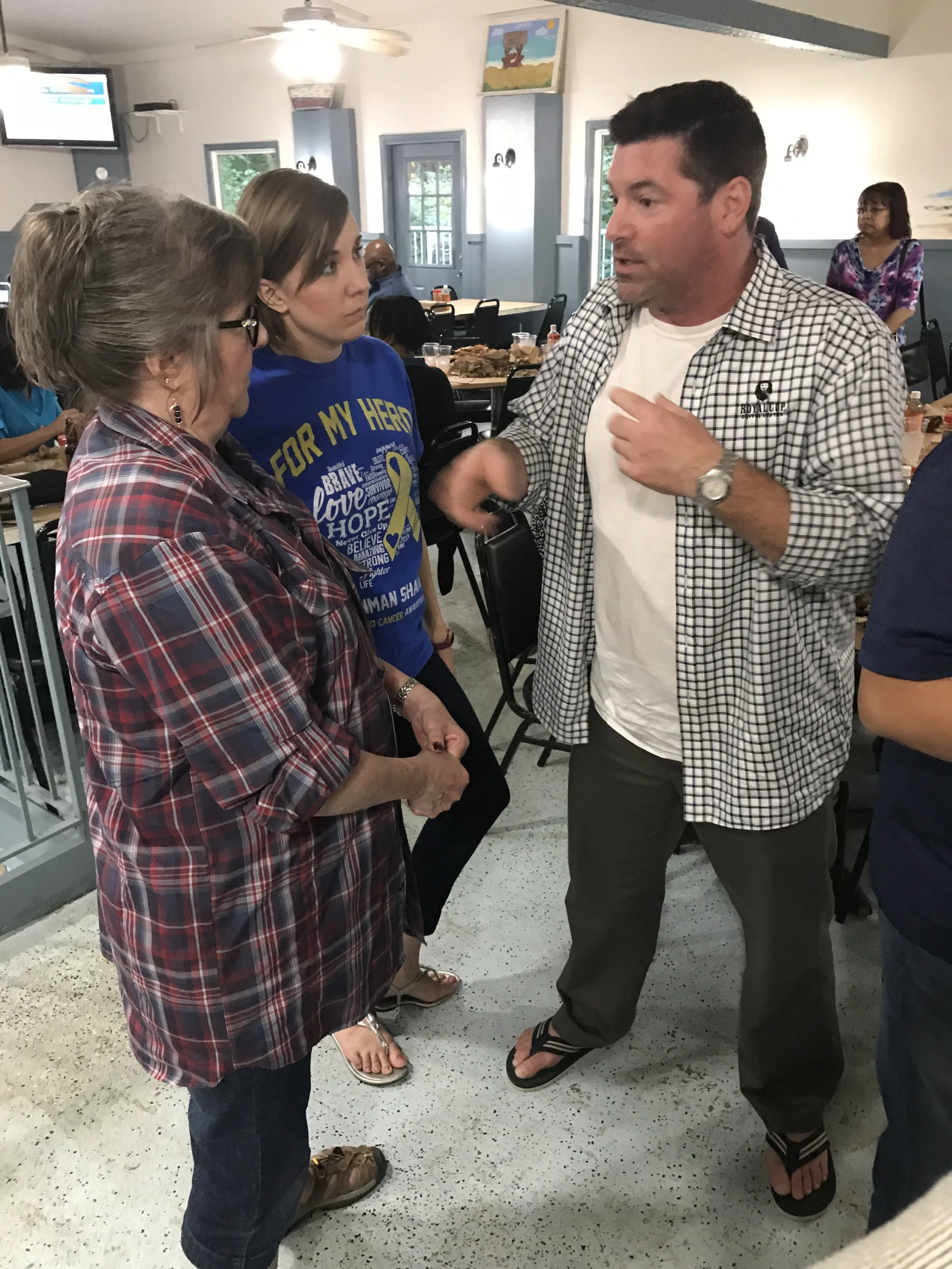 Three people engaged in conversation inside a restaurant or cafe. An older woman with glasses, a younger woman in a blue shirt, and a man wearing a checkered shirt.