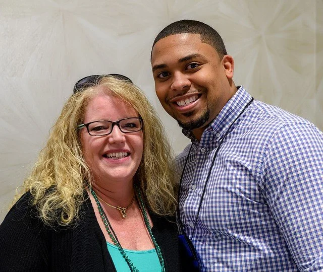 A woman with blonde curly hair, glasses, and a black top, smiling next to a young man with a short haircut and a checkered shirt, both smiling at the camera in an indoor setting.