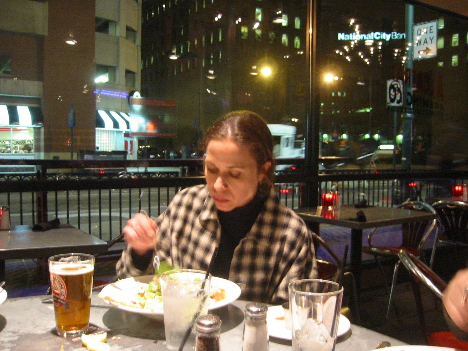 A woman with shoulder-length brown hair wearing a black turtleneck and checkered shirt, sitting at a restaurant table at night, with a city street and illuminated buildings, including a sign for National City Bank, visible through a large window behi