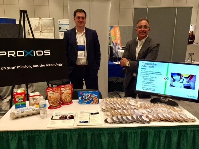 Two men standing behind a display table at a conference, with promotional materials, food samples, and a flat-screen monitor showing a presentation.