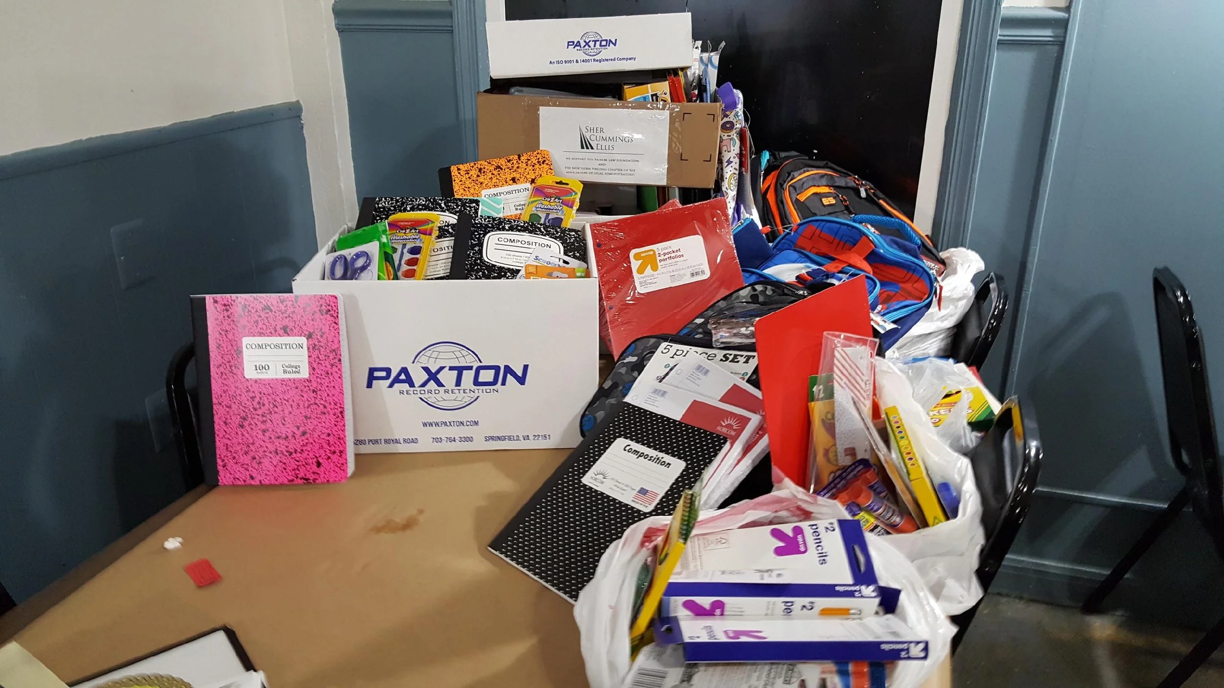 A table cluttered with school supplies including notebooks, pens, markers, glue, and a backpack, with boxes labeled for office and school use in a room with blue and gray walls.