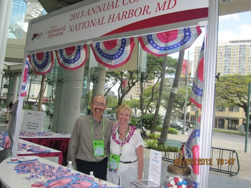 Two people standing inside a patriotic decorated booth at the 2013 Annual Conference & Exposition in National Harbor, MD, on April 25, 2012. The booth is decorated with red, white, and blue bunting.