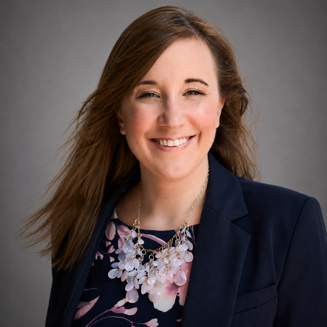 A professional woman with light skin and long brown hair, smiling, wearing a navy blazer, floral blouse, and a statement necklace, against a gray background.
