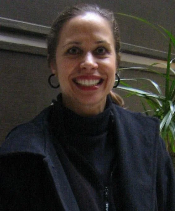 A woman smiling indoors, wearing a black zip-up jacket and hoop earrings, with a potted plant and a gray wall in the background.