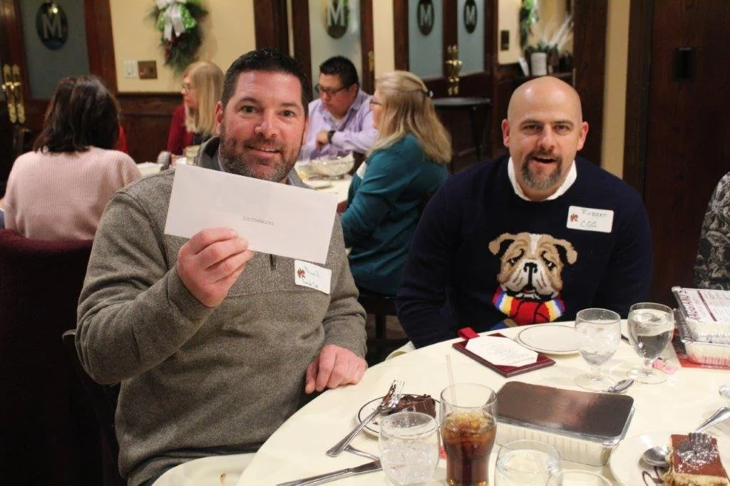 Two men sitting at a banquet table with others in the background, one holding a white envelope, in a decorated restaurant setting.
