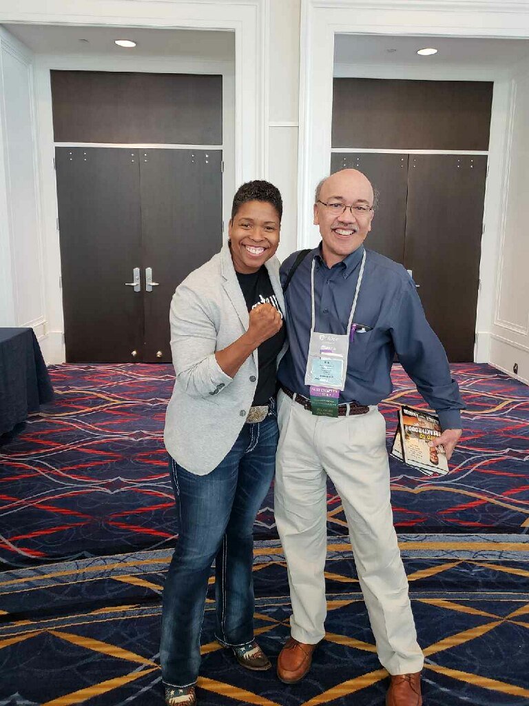 A woman and a man standing together in a conference room, smiling at the camera. The woman is wearing a gray blazer and jeans, and the man is wearing a blue shirt and light-colored pants, holding a book.