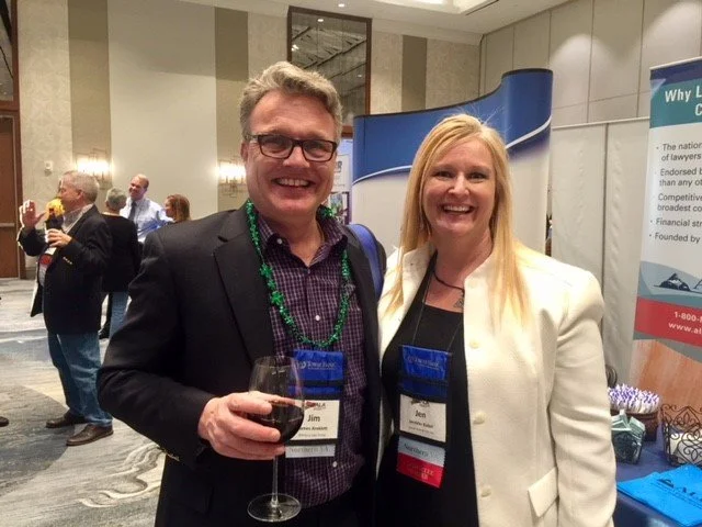 A man and woman smiling at a conference, holding wine glasses. The man wears glasses and a dark blazer, and the woman wears a white blazer. They have name tags and are standing in front of a blue and white booth.