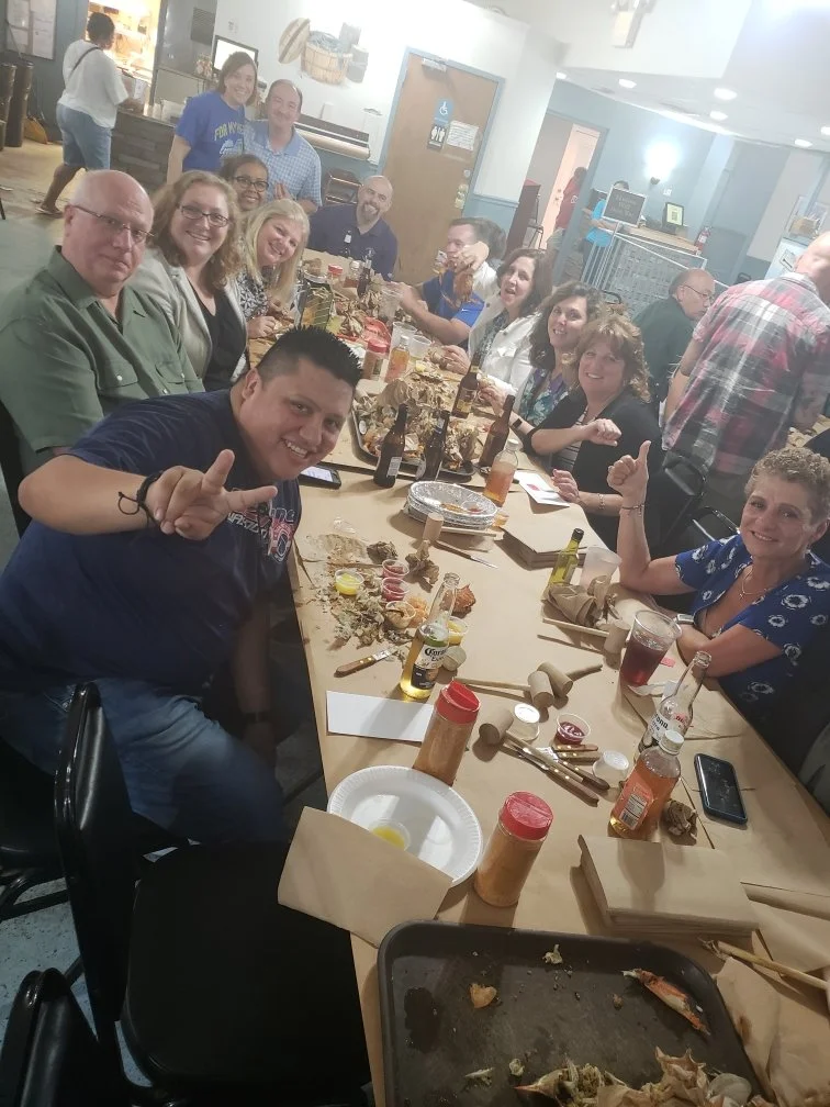A group of people seated at a long table, enjoying a meal together in a restaurant. They are smiling, some giving thumbs up, with food, drinks, and condiments on the table.