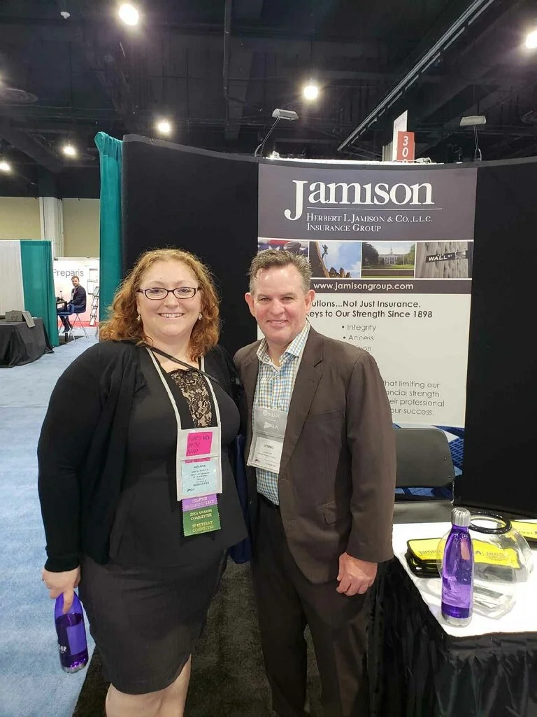 A woman and a man standing together at a trade show booth for Jamison insurance group, smiling and facing the camera.