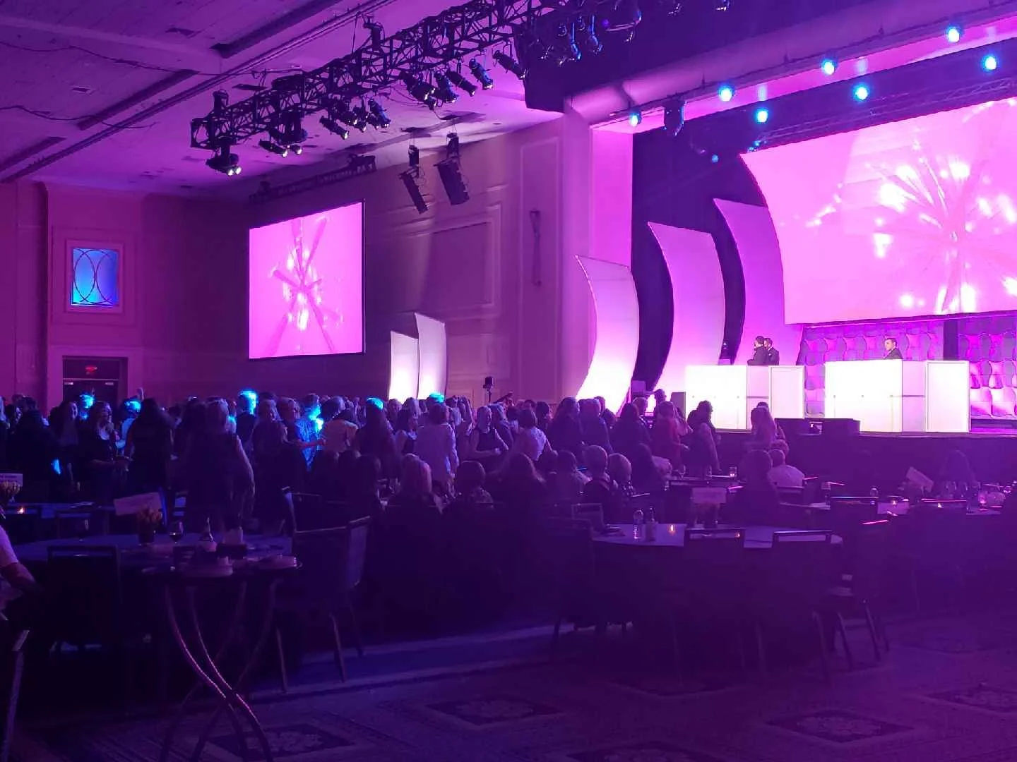 A large conference room with a stage bathed in purple and pink lighting, featuring two large screens displaying abstract visuals, and a crowd of attendees watching the event.