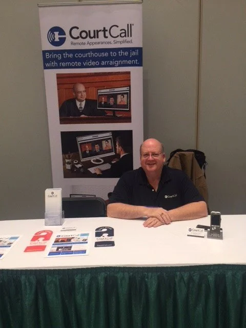 A man sitting at a table with promotional materials and electronic devices, behind a banner for CourtCall, a remote court appearance service.