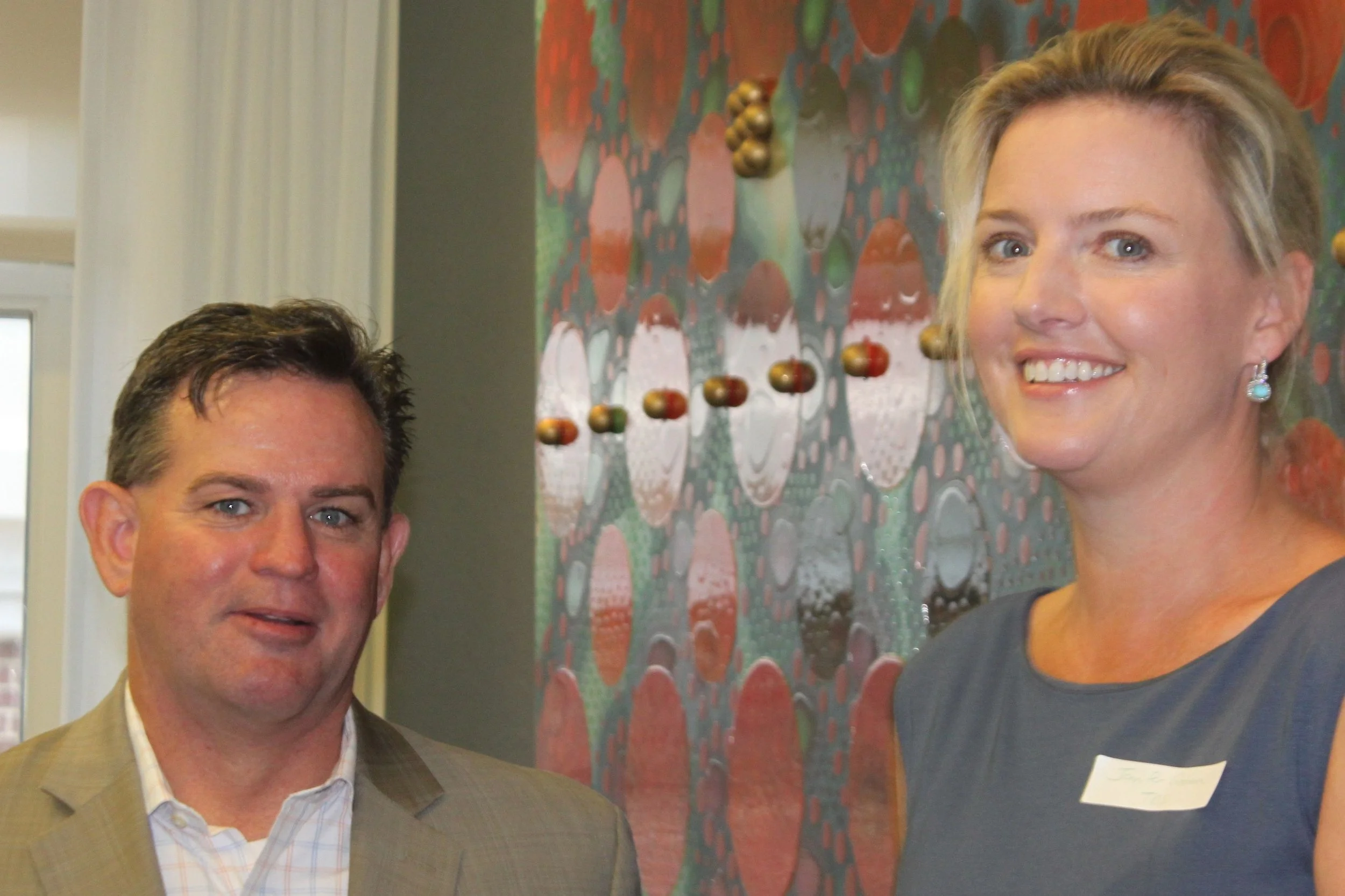 A man and a woman standing in front of a colorful abstract wall art, smiling at the camera. The man wears a beige blazer and white shirt, and the woman wears earrings and a dark top.