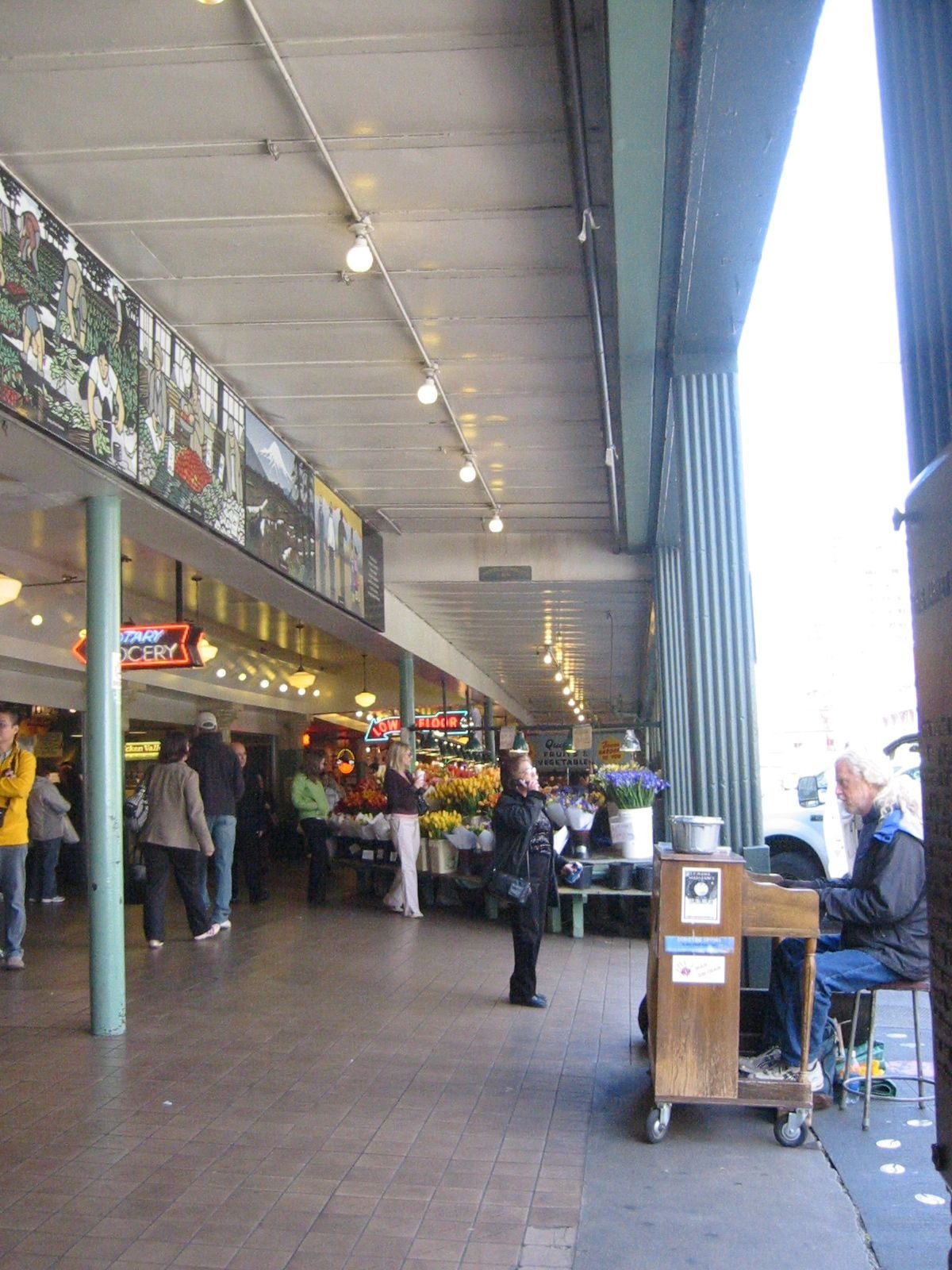 A busy marketplace or shopping area with a flower stand, people shopping and walking, and a man playing piano with a cart nearby, under a covered walkway with lights and signage.