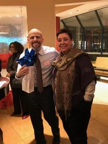 Two people standing indoors, smiling, one holding a gift bag.