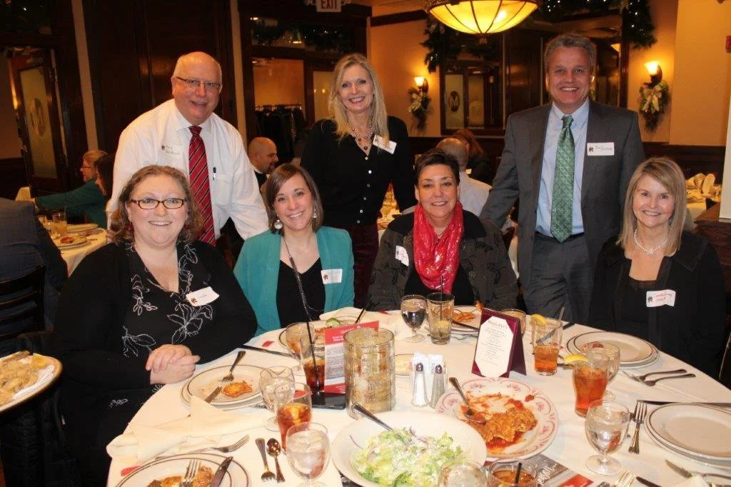 Group of seven people, sitting and standing around a round dining table, smiling, at a formal event in a restaurant or banquet hall decorated with Christmas ornaments.