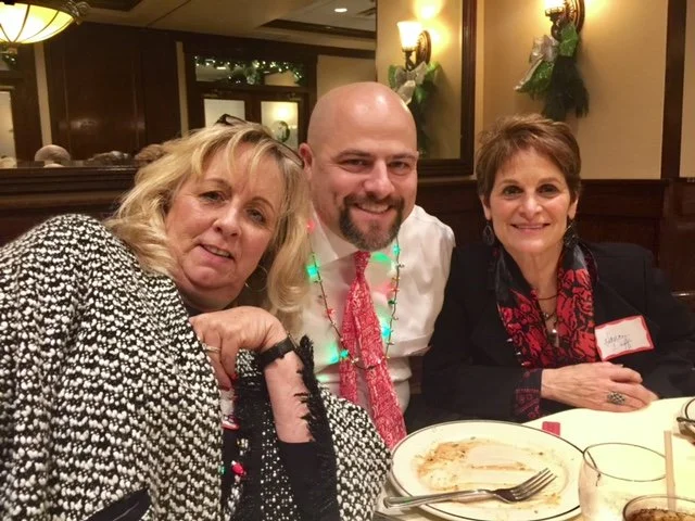 Three people sitting at a restaurant table during Christmas, smiling and dressed in festive attire, with decorations and dinnerware visible.