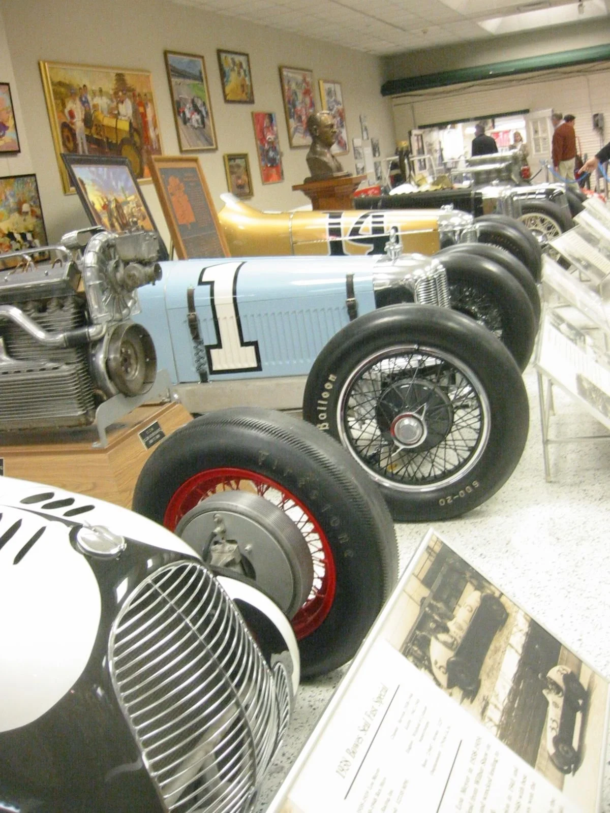 Collection of vintage racing cars displayed in a museum, with framed artwork and a bust sculpture on the wall, and informational plaques in the background.