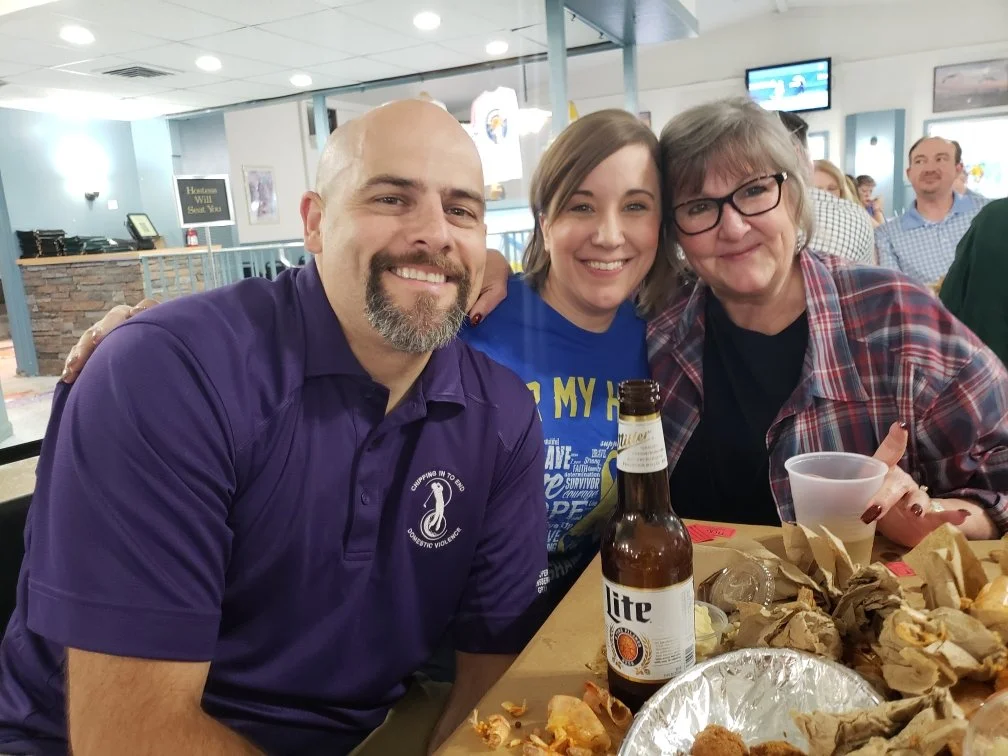 Three people sitting at a table, smiling. A man in a purple shirt, woman in a blue shirt, and woman with glasses and a plaid shirt. There are food and drinks on the table.