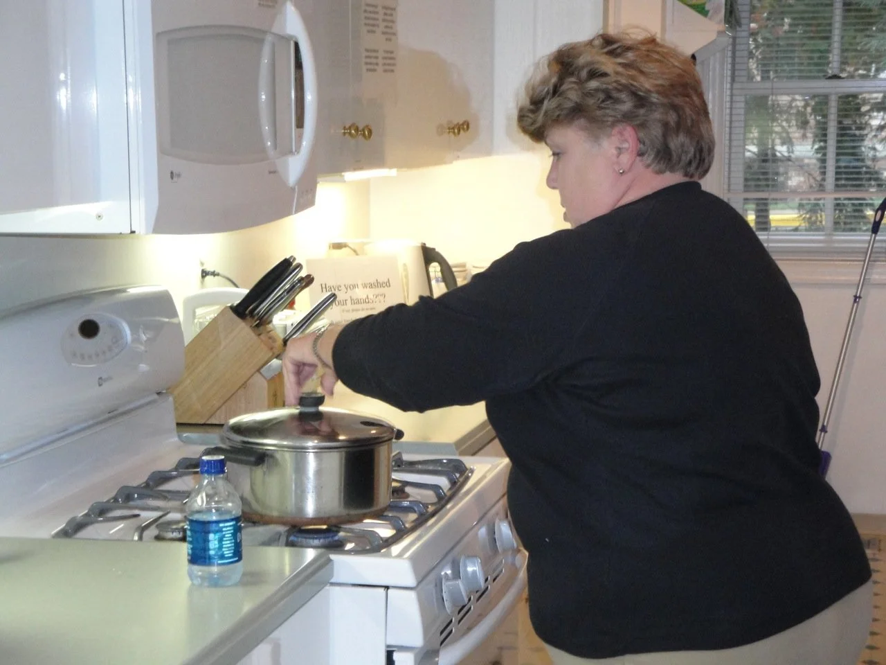 A woman cooking on a stove in a kitchen, lifting the lid off a pot with a water bottle on the counter nearby.