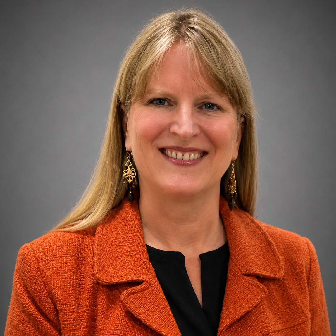 Portrait of a woman with blonde hair, wearing earrings and an orange blazer, smiling against a gray background.