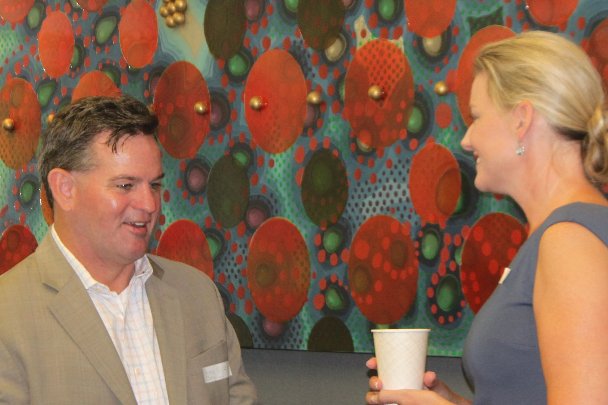 A man in a beige suit and a woman with blonde hair are talking at an event, with a colorful abstract art piece on the wall behind them. The woman is holding a white paper cup.